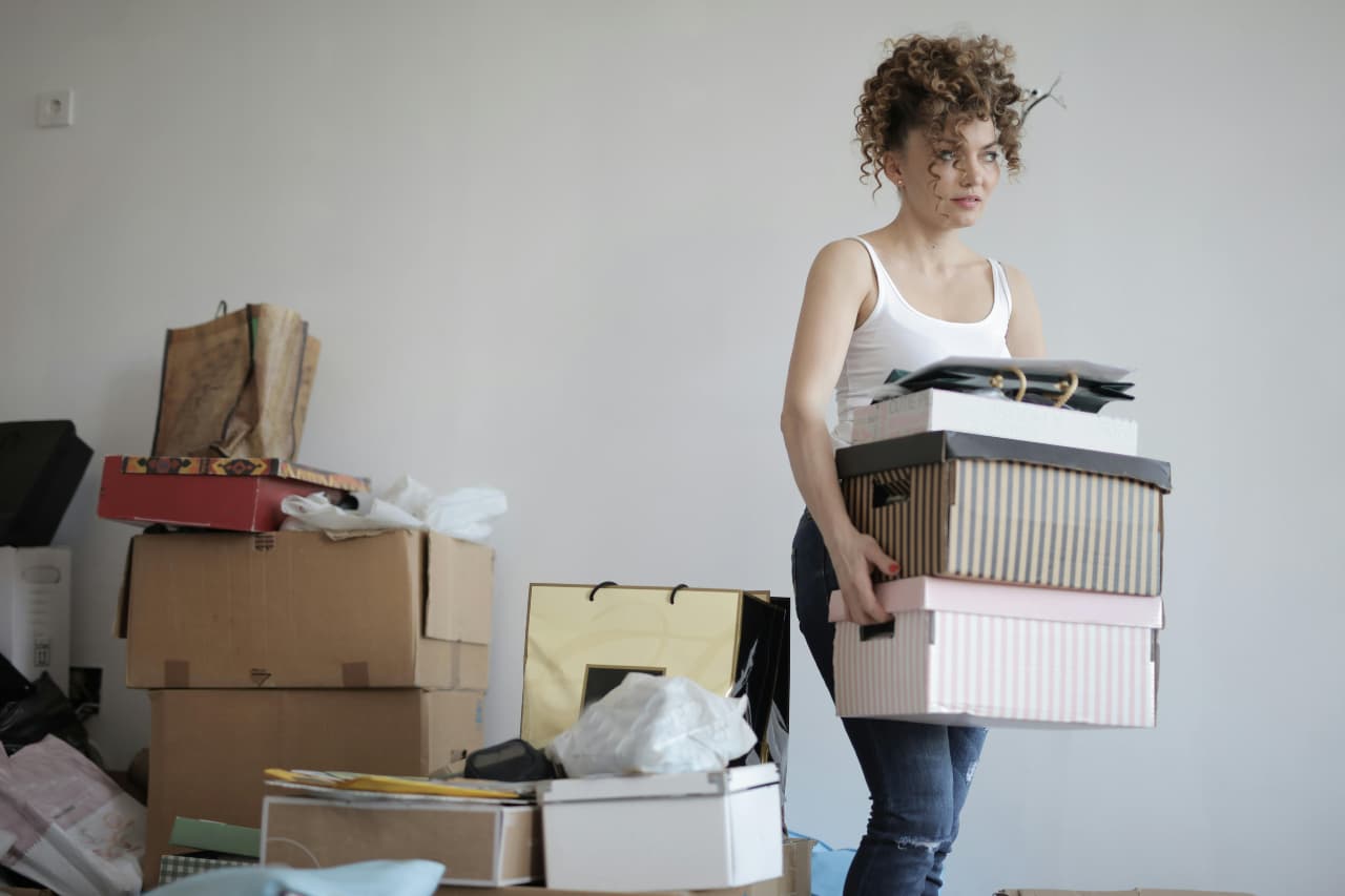 Woman carrying stacked boxes in a room filled with packed items.